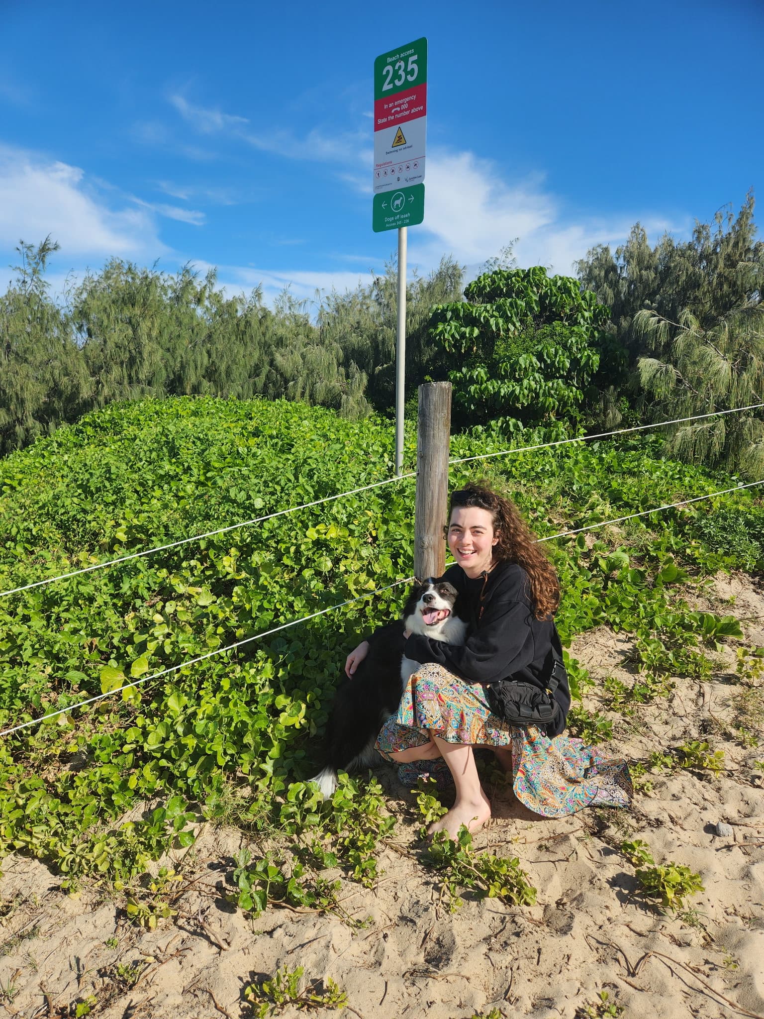 Smiling woman in a patterned skirt hugs a border collie on a sandy beach path.